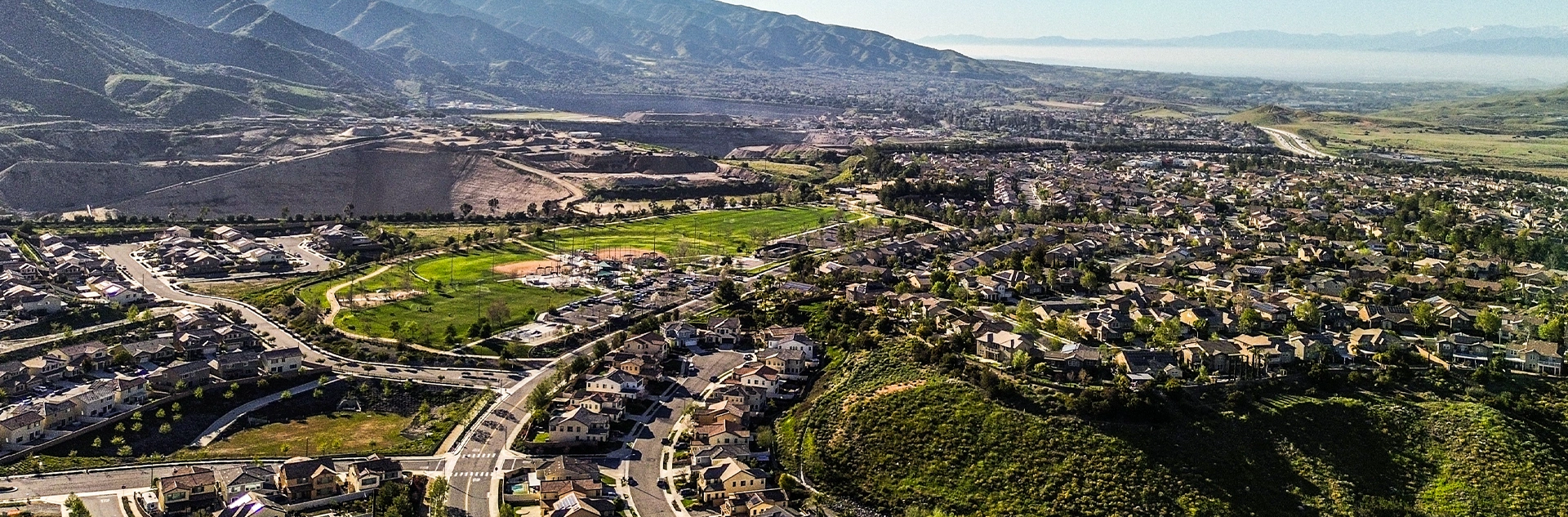 Aerial view of a suburban neighborhood with houses, winding roads, a green park, and mountains in the background under a clear sky.