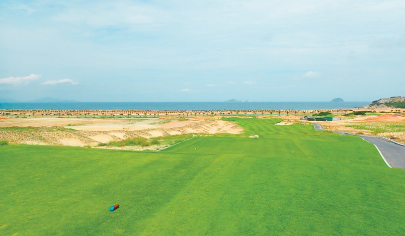 A wide, green golf course stretches toward the sea under a blue sky. Sandy patches and a small paved path are visible, with distant hills and calm ocean water in the background.