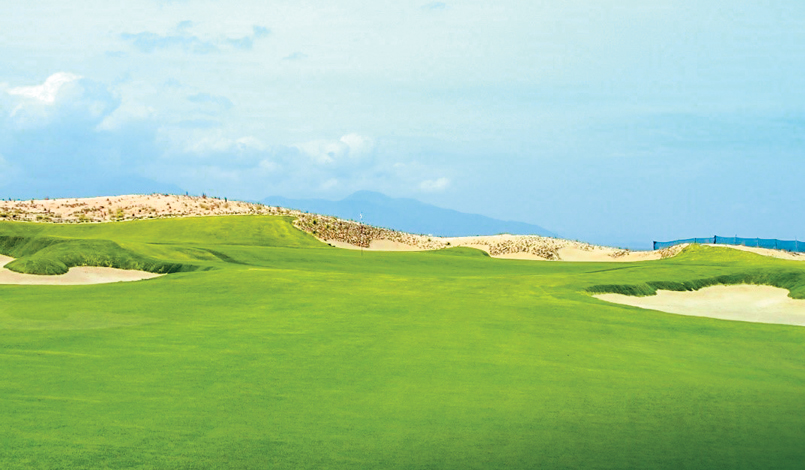 Wide view of a golf course with bright green grass, sand bunkers, and a distant sandy hill under a cloudy blue sky. Mountains are faintly visible in the far background.