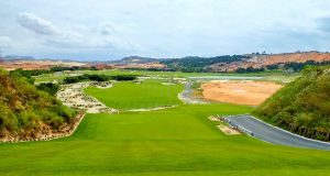 A scenic golf course with green fairways, sand bunkers, and rolling hills under a partly cloudy sky, surrounded by natural vegetation and dirt paths.