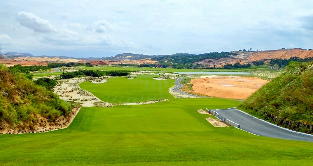 A scenic golf course with green fairways, sand bunkers, and rolling hills under a partly cloudy sky, surrounded by natural vegetation and dirt paths.