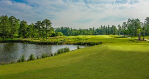 Lush golf course with a pond on the left, green fairways, scattered trees, and a partly cloudy sky in the background.