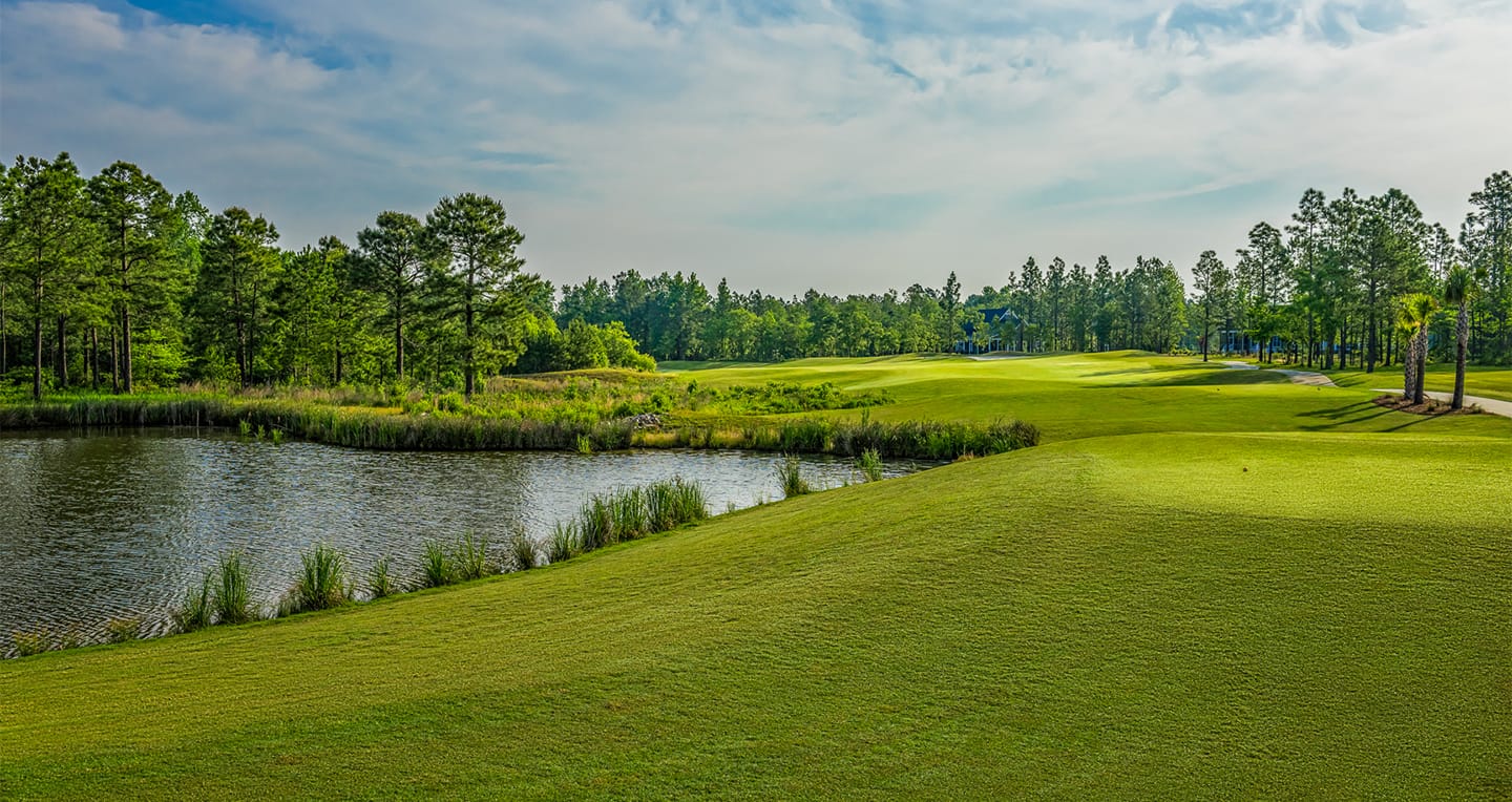 Lush golf course with a pond on the left, green fairways, scattered trees, and a partly cloudy sky in the background.