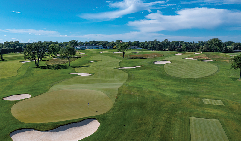 A wide view of a golf course with green fairways, several sand bunkers, trees, and a blue sky with clouds in the background.