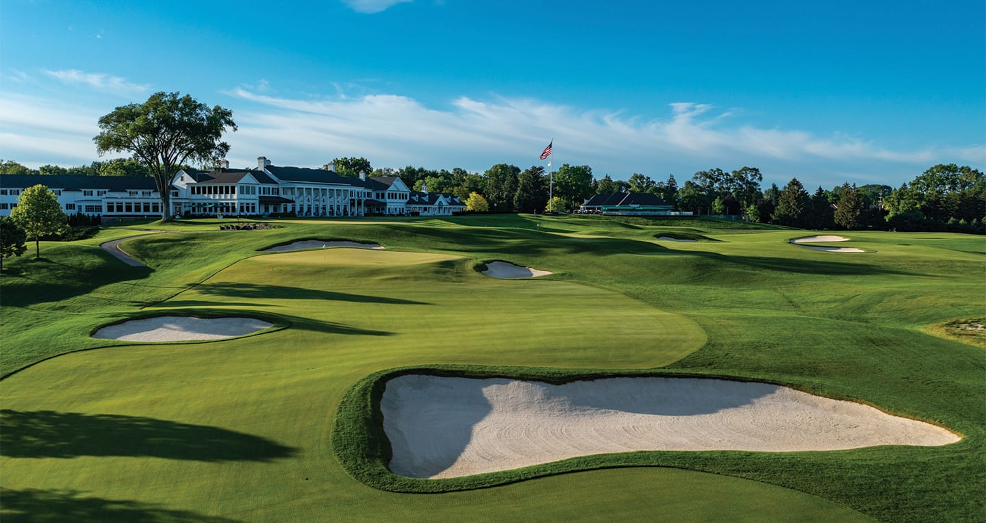 A lush green golf course with sand bunkers and a large clubhouse in the background under a clear blue sky.