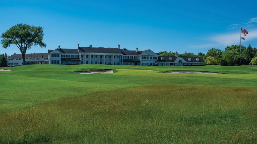 A large clubhouse sits behind a lush green golf course with sand bunkers, a tall tree, and an American flag waving under a clear blue sky.