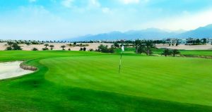 A golf green with a flagstick, surrounded by sand traps and palm trees, with mountains and buildings in the background under a partly cloudy sky.