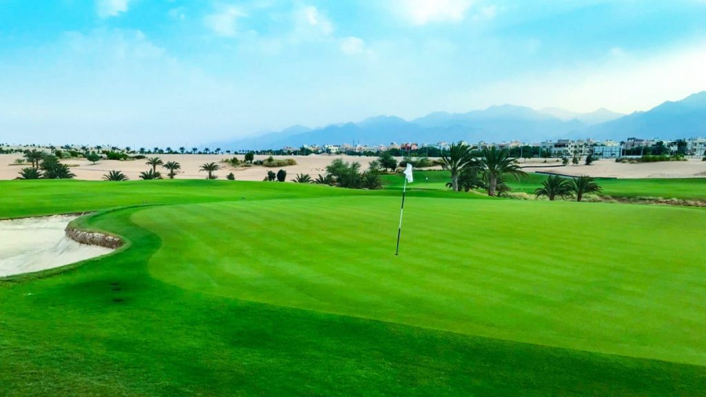 A golf course with a putting green, a sand bunker to the left, palm trees, and distant mountains under a partly cloudy sky.