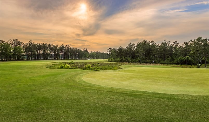 A golf course with neatly trimmed grass, a sand bunker, and trees lining the horizon under a partly cloudy sky at sunset.