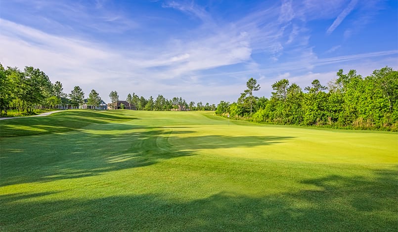 A wide, green golf course fairway under a blue sky with light clouds, surrounded by trees and a few buildings in the distance.