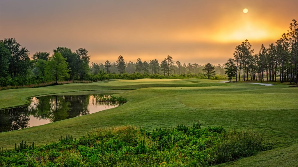 A scenic golf course at sunrise, with sunlight reflecting on a pond, lush green grass, and trees lining the horizon under a partly cloudy sky.