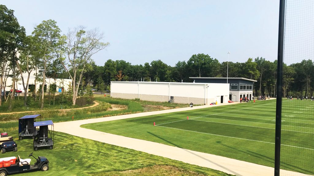 A green sports practice field with white boundary lines, a paved path, utility vehicles parked nearby, and a modern building surrounded by trees in the background under a clear sky.