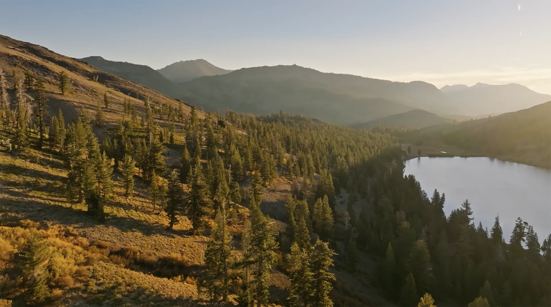 Sunlight shines over a forested mountain landscape with a reflective lake on the right, gentle slopes, and distant peaks under a clear sky.