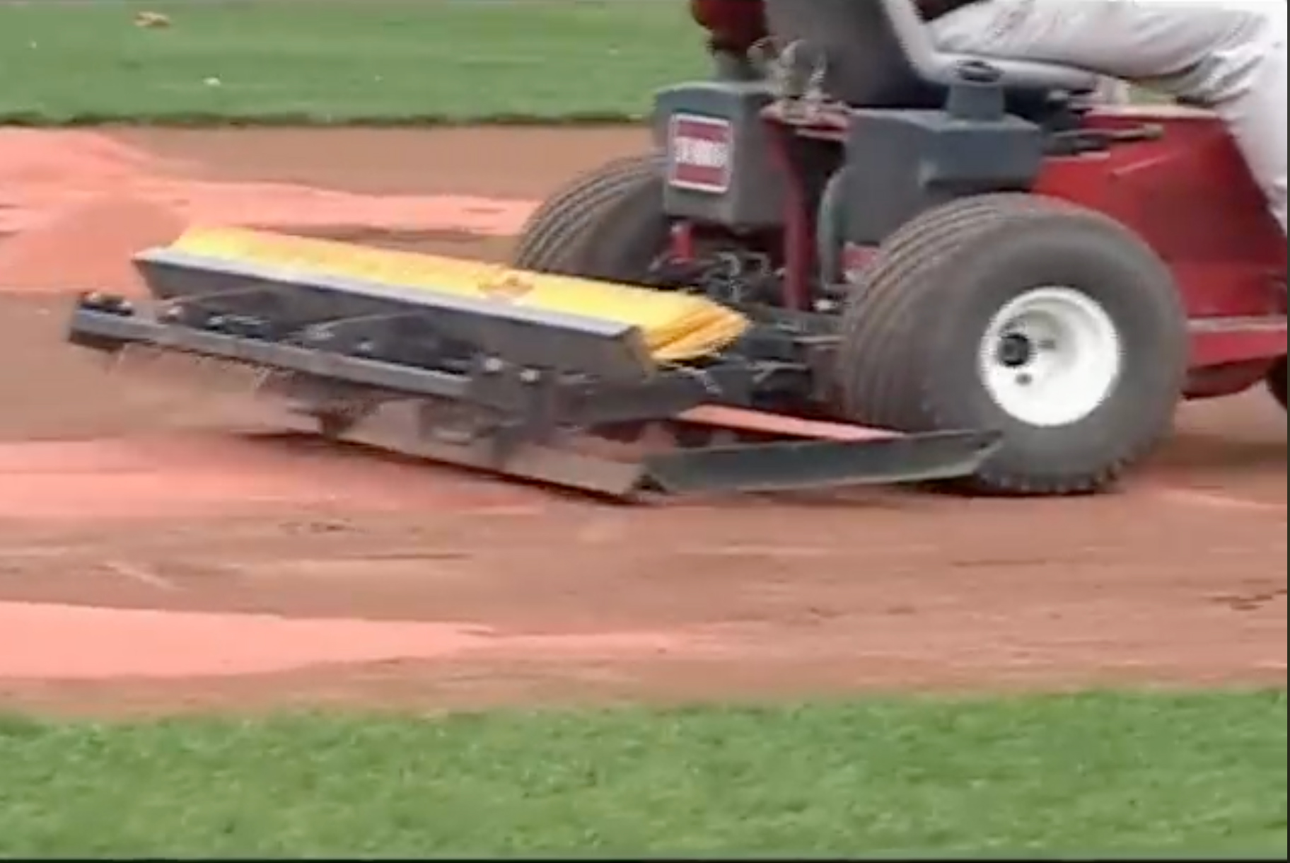 A person drives a red field-grooming machine over a baseball or softball infield, smoothing the dirt near the grass edge.