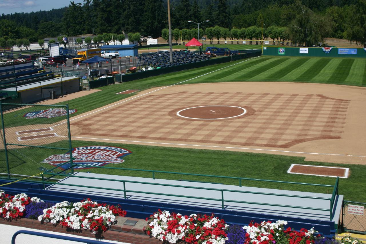 A well-maintained baseball field with checkerboard grass patterns, empty stands, and vibrant flower beds in the foreground on a sunny day.