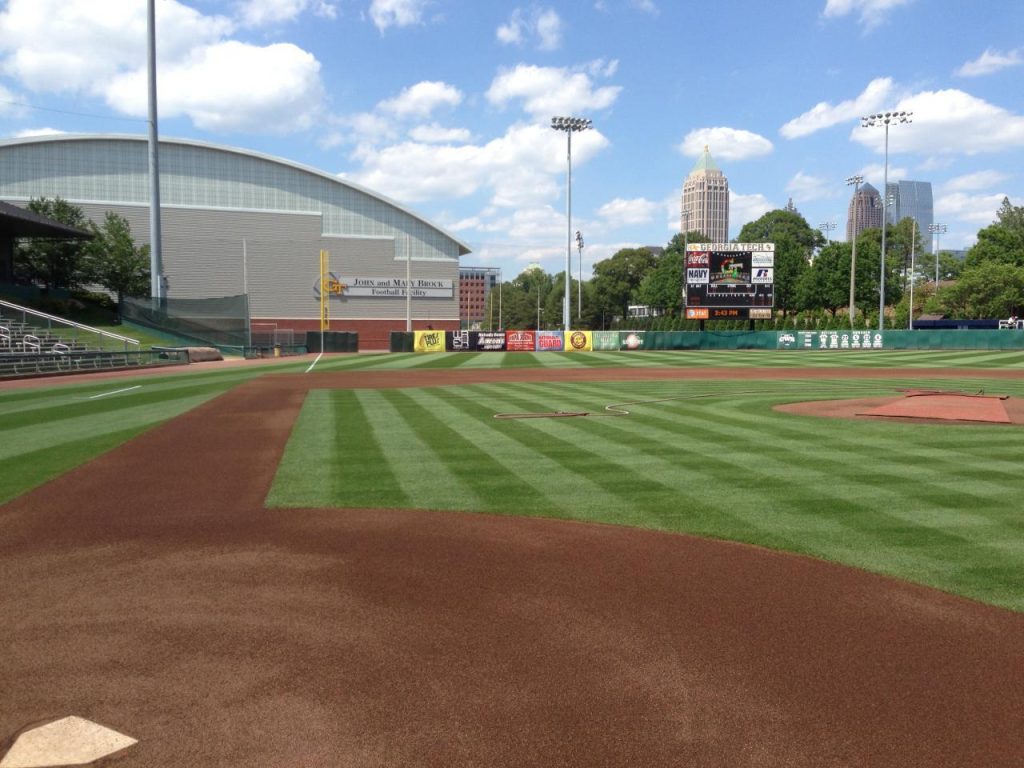 A well-maintained baseball field with green grass, brown dirt, and scoreboard in the background under a blue sky with clouds. Buildings and trees surround the stadium.