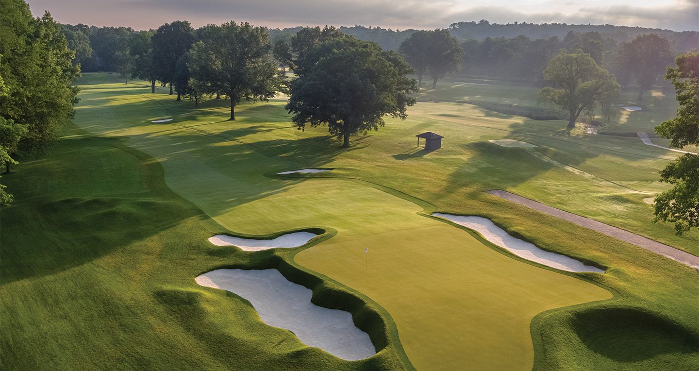 A golf course at sunrise with manicured greens, white sand bunkers, large trees, and long shadows cast across the fairway under a partly cloudy sky.