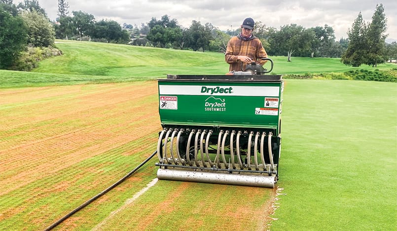 A person operates a DryJect machine on a golf course, aerating and injecting sand into the turf. The machine leaves a strip of treated grass next to untreated green grass.