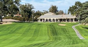 A golf course with neatly mowed green grass leads to a clubhouse surrounded by trees under a blue sky. A paved path curves along the right side of the fairway.