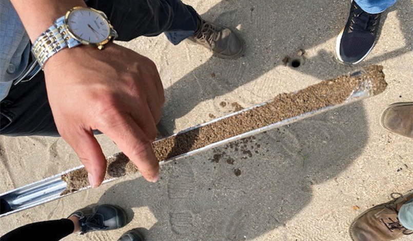 A persons hand holds a metal soil core sampler filled with sand, surrounded by several people standing on sandy ground. Only their hands, feet, and shoes are visible.