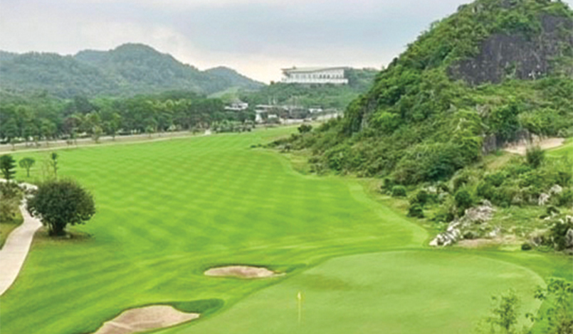 A lush green golf course with sand bunkers, rolling hills, trees, and a large building in the distance under a cloudy sky.