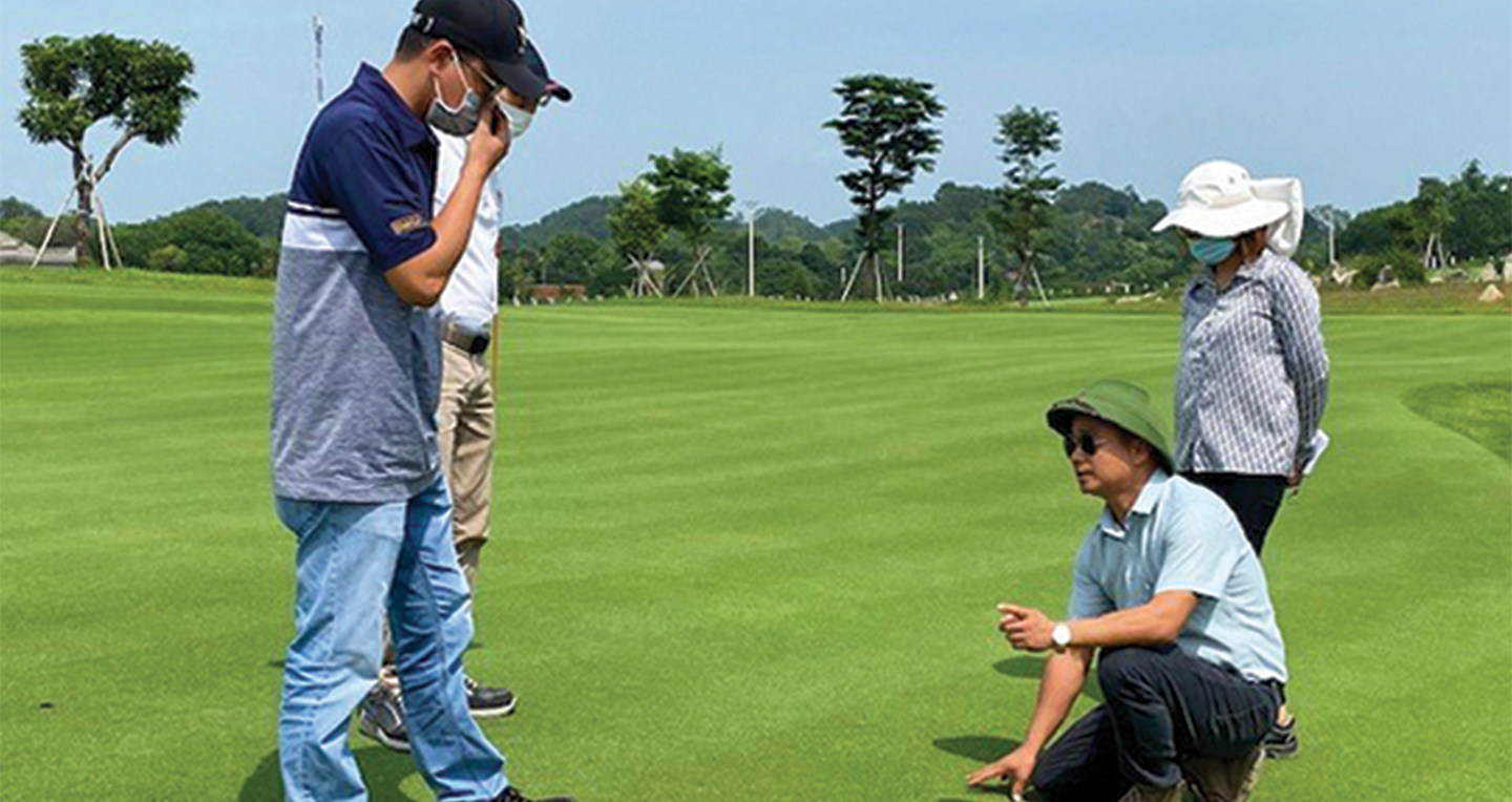 Four people on a golf course, two standing, one kneeling and pointing at the grass, all wearing hats and masks, with trees and a clear sky in the background.