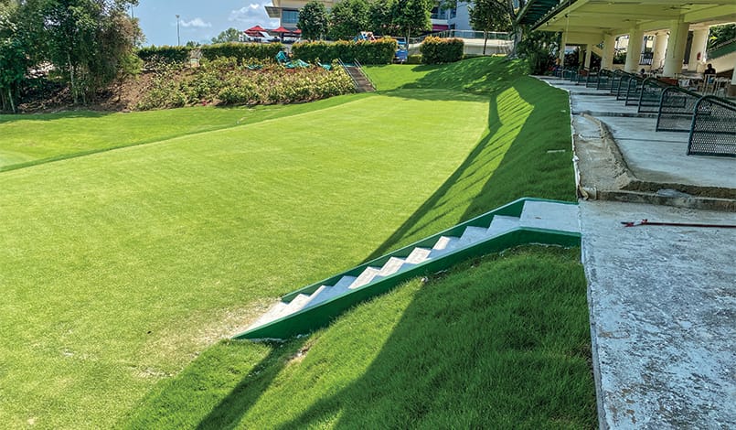 A green golf driving range with a row of practice bays on the right, green grass on the left, and a small staircase leading down to the field. Some trees and a building are visible in the background.