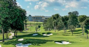 A lush golf course with green fairways, sand bunkers, and trees, with a modern clubhouse building in the background under a partly cloudy sky.