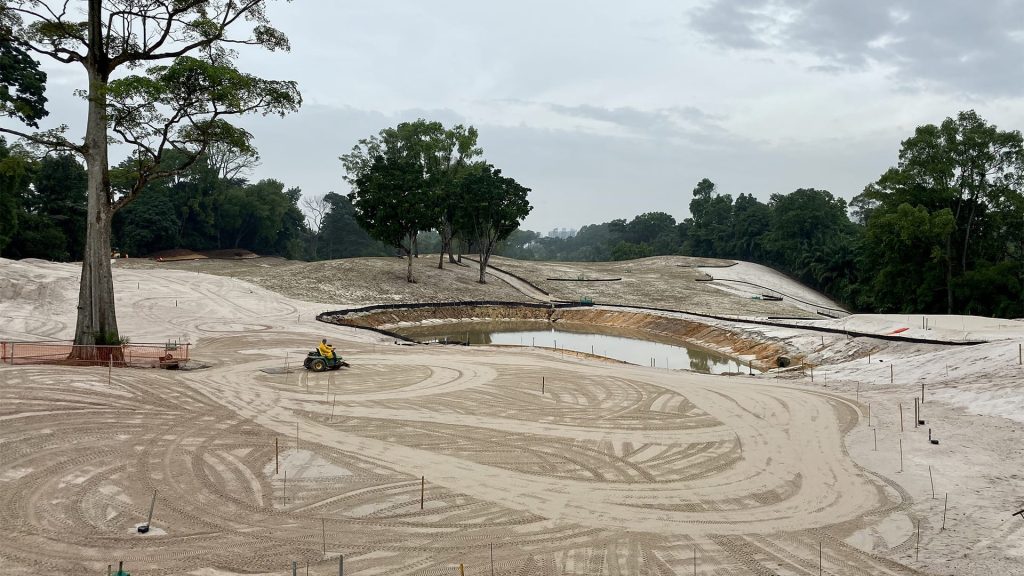 A construction site with sandy ground, tire tracks, a small pond, scattered trees, and a worker operating a vehicle under a cloudy sky.