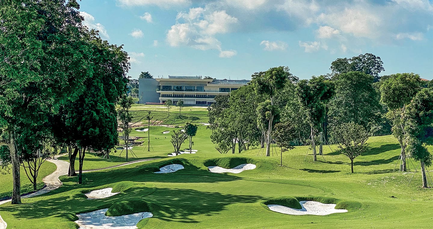 A lush golf course with green fairways, sand bunkers, and trees, with a modern clubhouse building in the background under a partly cloudy sky.