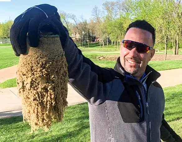 A man wearing sunglasses and gloves holds up a clump of wet, sandy material outdoors on a sunny day in a park with green grass and trees in the background.