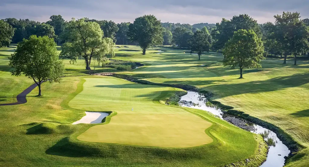 A lush golf course with manicured greens, sand bunker, winding creek, and trees under a cloudy sky. The scene is peaceful and well-maintained.