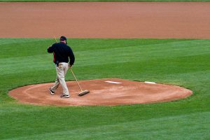 A groundskeeper sweeps the pitchers mound on a baseball field, preparing the area before a game. The grass surrounding the mound is neatly trimmed and the infield is well-maintained.
