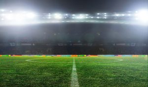 A well-lit soccer stadium at night, viewed from midfield, with bright lights shining over the empty green pitch and blurred audience seating in the background.