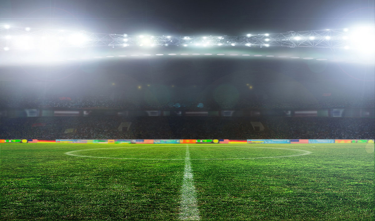 A well-lit soccer stadium at night, viewed from midfield, with bright lights shining over the empty green pitch and blurred audience seating in the background.