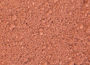 Close-up of loose, reddish-brown soil or sand, showing a rough, grainy texture with small clumps and particles throughout the surface.