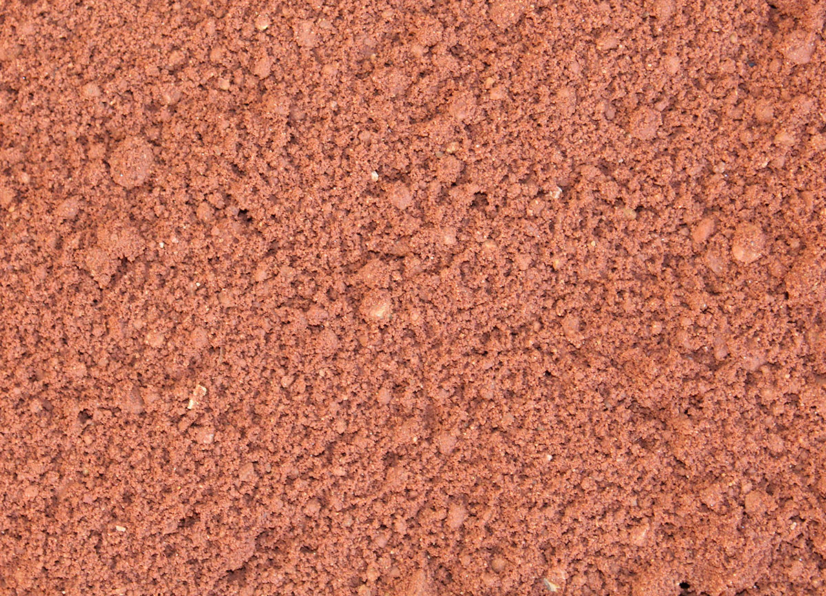 Close-up of loose, reddish-brown soil or sand, showing a rough, grainy texture with small clumps and particles throughout the surface.