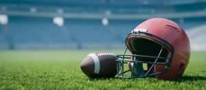 A football and a red helmet rest on a green field in an empty stadium with blurred seating in the background.