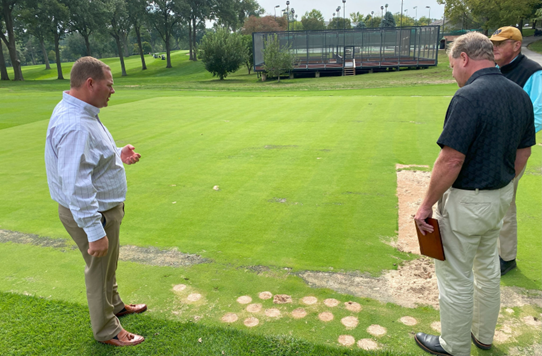 Three men stand on a golf course, examining patches of grass and dirt spots. One man holds a notebook. Trees and a fenced area are visible in the background.