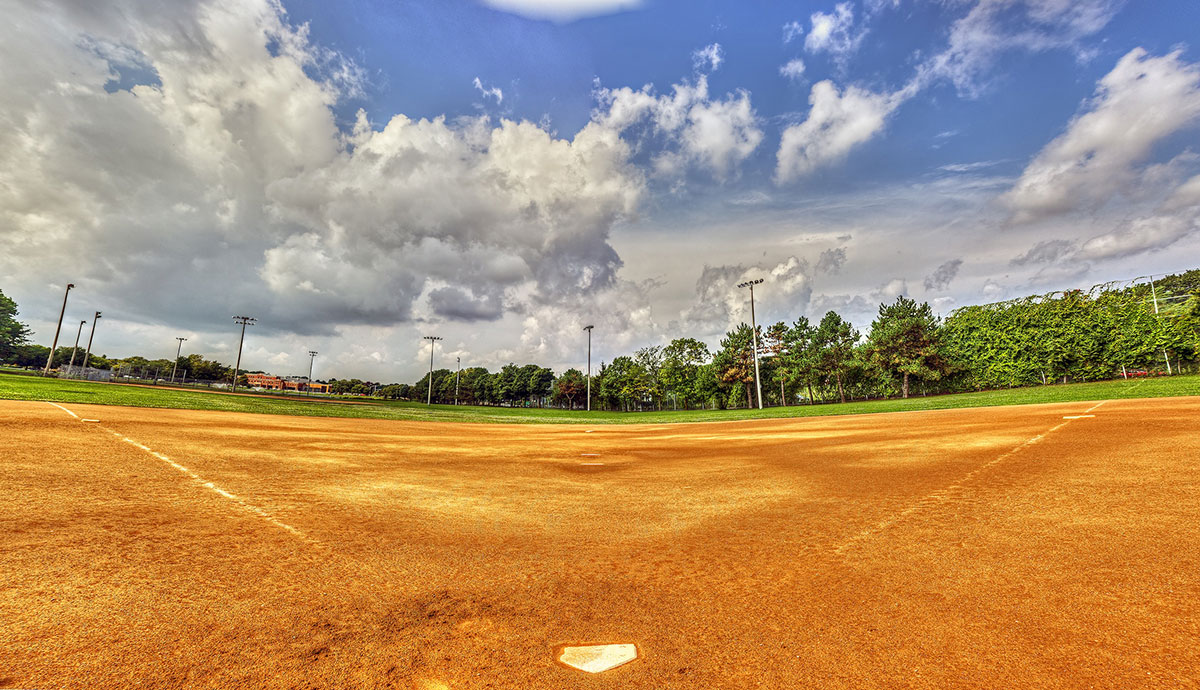 Empty baseball field with a clear home plate, orange-brown infield dirt, green grass outfield, and tall trees in the background under a partly cloudy blue sky.