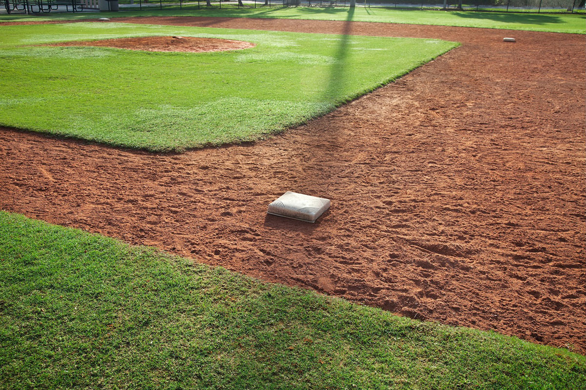 A close-up view of a baseball field showing a base, the infield dirt, the pitchers mound, and green grass on a sunny day.