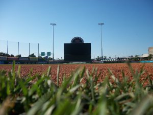 Low-angle view from the infield of a baseball stadium showing grass, dirt, and an empty scoreboard under a clear blue sky. Stadium lights and some signs are visible in the background.