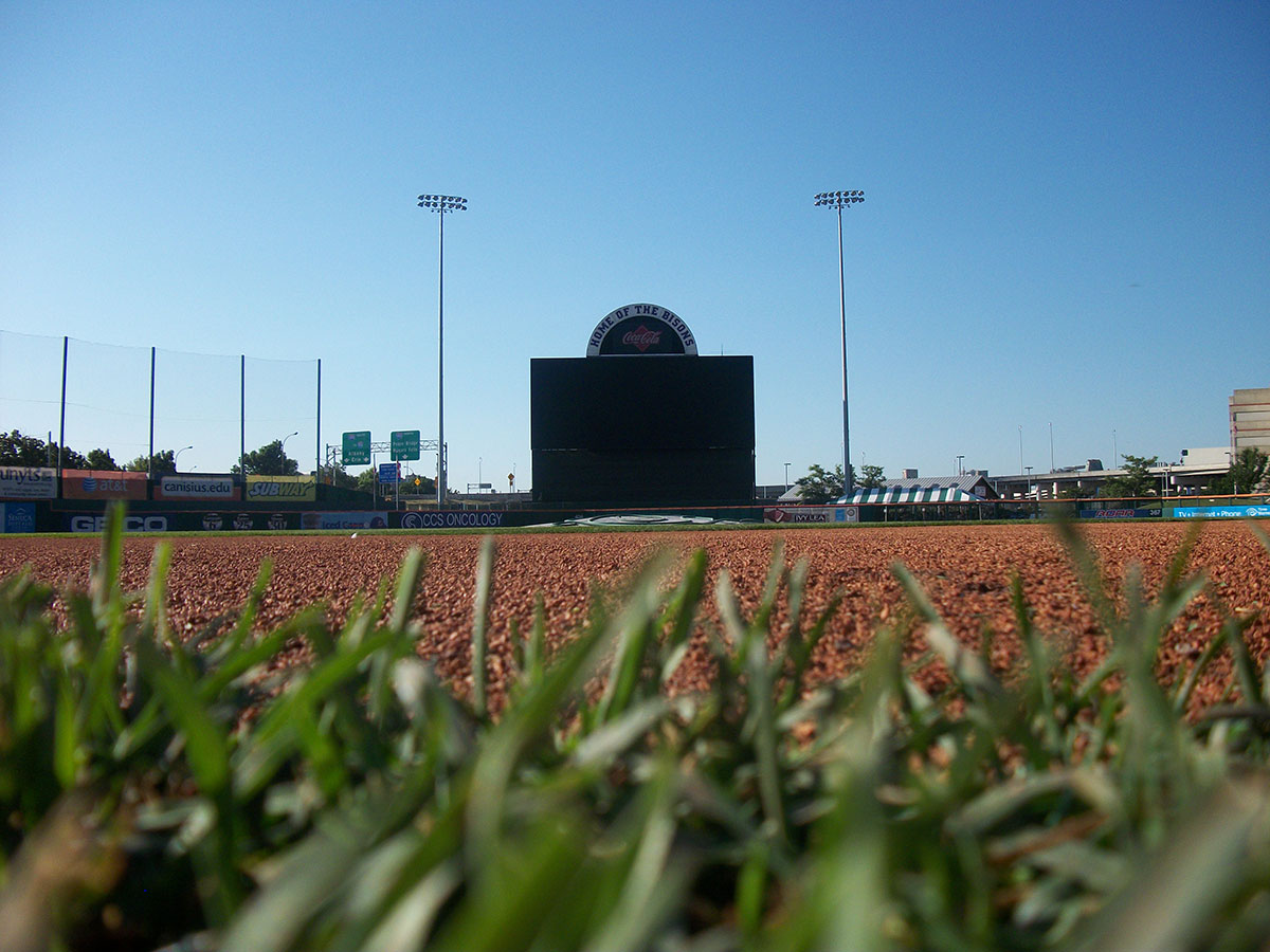 Low-angle view from the infield of a baseball stadium showing grass, dirt, and an empty scoreboard under a clear blue sky. Stadium lights and some signs are visible in the background.