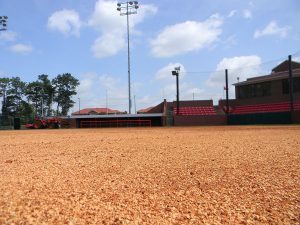 A low-angle view of an empty baseball or softball field with red gravel, a dugout, bleachers, a tractor, and lights under a partly cloudy sky.