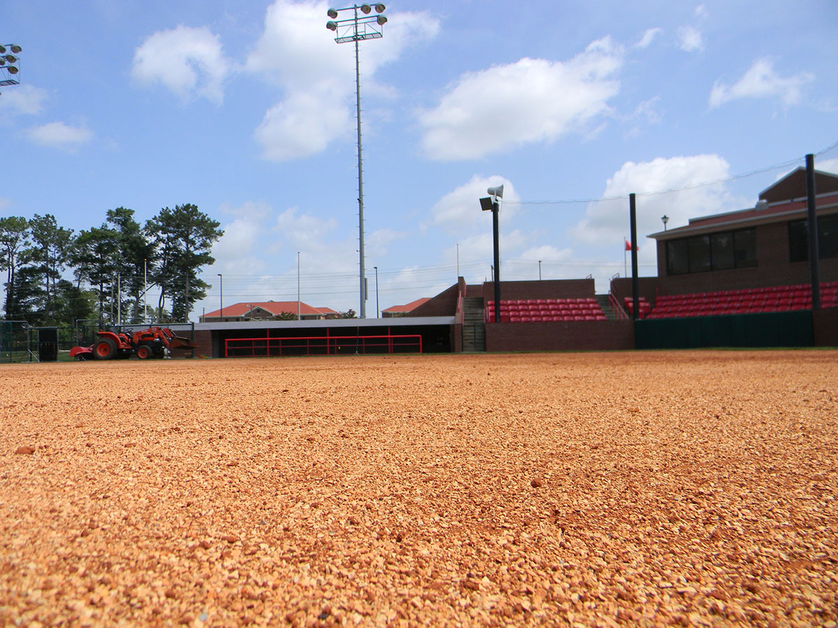A low-angle view of an empty baseball or softball field with red gravel, a dugout, bleachers, a tractor, and lights under a partly cloudy sky.