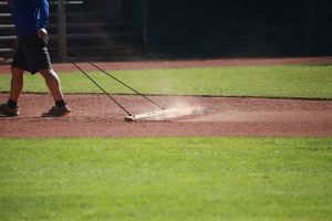 Person dragging a dirt rake to groom the infield of a baseball field, kicking up dust as they walk, with green grass and a chain-link fence visible in the background.