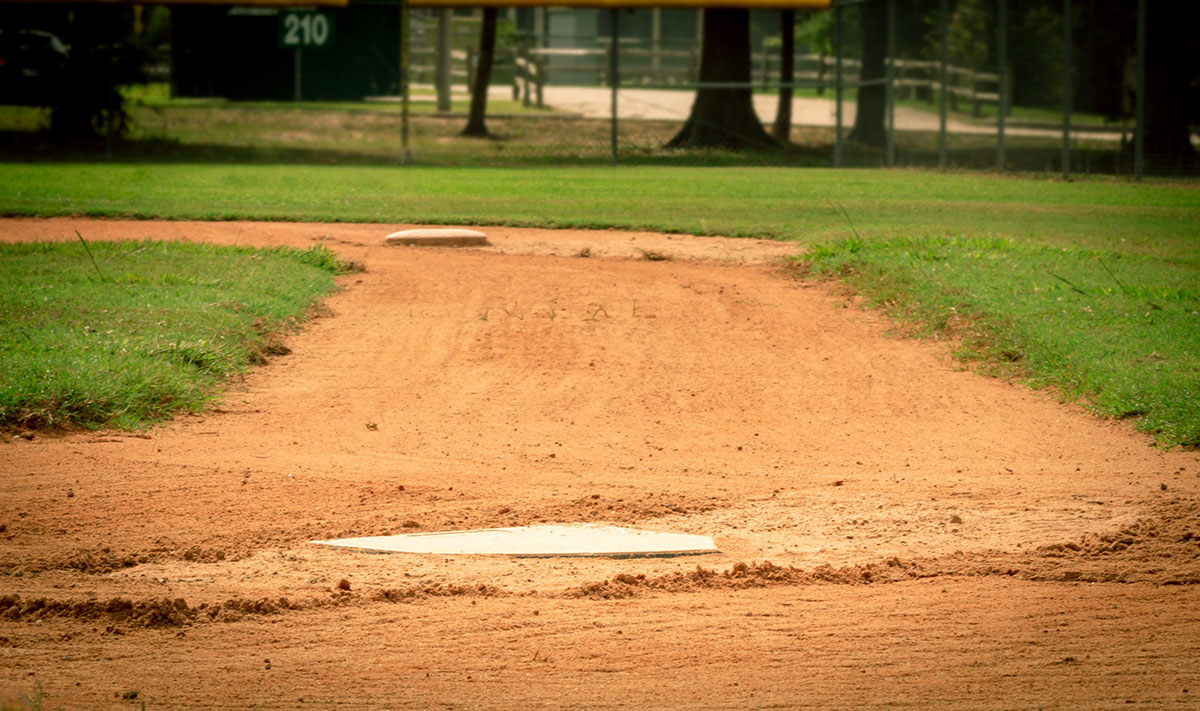 A close-up view of a baseball field shows home plate in the foreground and second base in the distance, with green grass and a fence in the background.