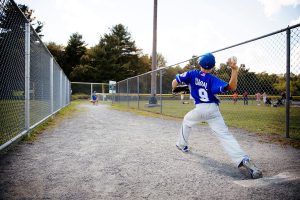 A young baseball player in a blue jersey winds up to throw a pitch on a gravel path, aiming toward a catcher behind a chain-link fence on a sunny day.
