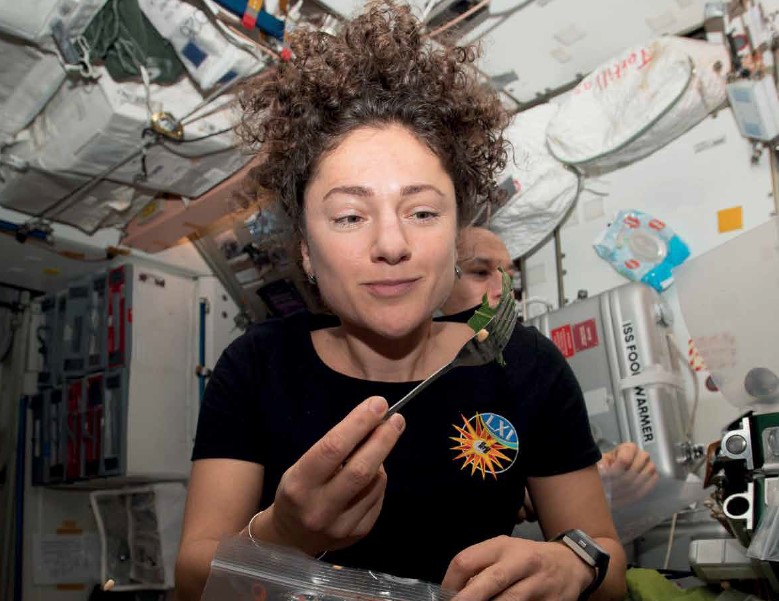 Astronaut in microgravity inspecting fresh leafy greens with a fork inside the International Space Station.