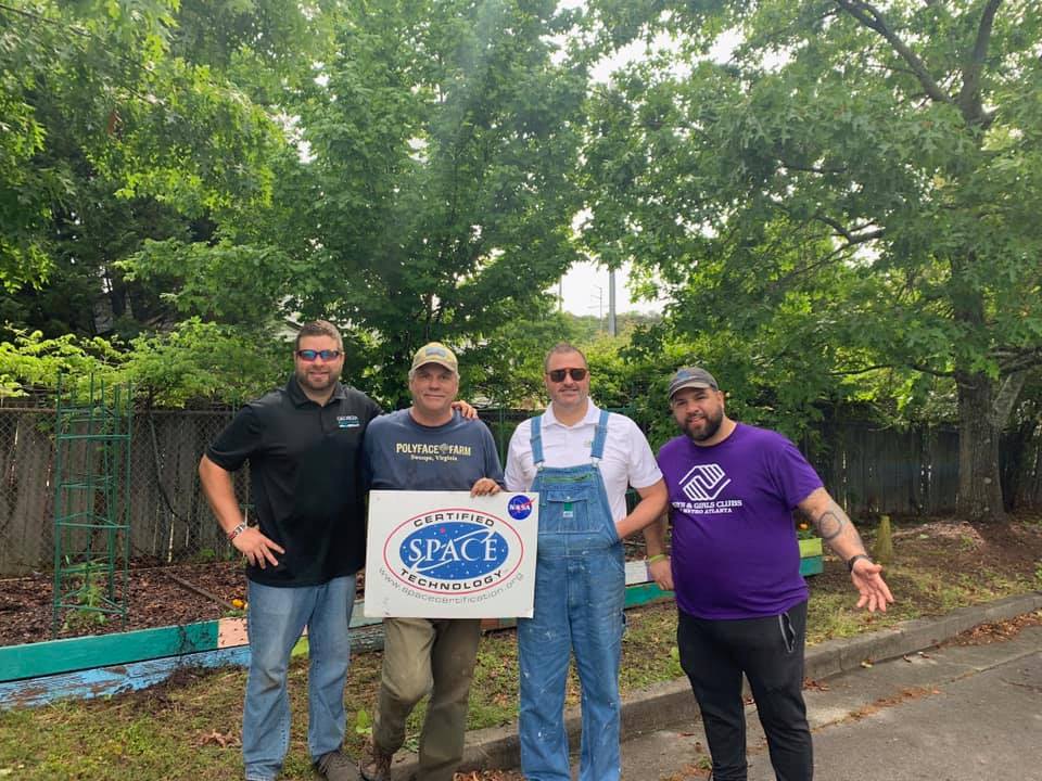 Four men pose outdoors in a garden holding a NASA “Certified Space Technology” sign.
